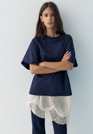 Giovane donna con capelli lunghi e scuri in piedi con le braccia incrociate, indossando una t-shirt oversize blu navy sovrapposta a una maglia bianca con bordi in pizzo e jeans scuri.