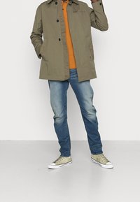 Giacca leggera verde con chiusura a bottoni, polo arancione, jeans blu e sneaker verde oliva. Outfit casual con colori a contrasto.