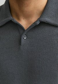 Primer plano del cuello de una persona que lleva una camiseta tipo polo de punto gris oscuro con un solo botón abotonado.