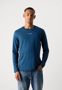 Jonge man in een blauw langemouwen Marc O'Polo shirt en blauwe spijkerbroek, staand met één hand in zijn zak tegen een effen achtergrond.
