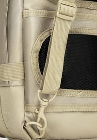 Detail béžového látkového batohu se strukturovanou síťovinou, nastavitelním popruhem a pevným klipovým uchycením.