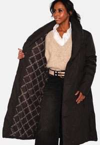 Lang svart dunjakke med høy krage. Innside har et quiltet mønster i mørkebrun med hvite detaljer. Foran synlige trykknapper.