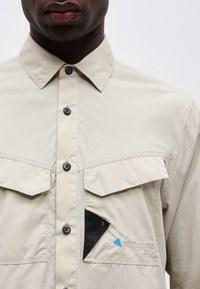 Άνδρας που φοράει μπεζ πουκάμισο με κουμπιά και δύο τσέπες στο στήθος, με το καπάκι μιας τσέπης να έχει σηκωθεί μερικώς αποκαλύπτοντας μαύρο υφασμάτινο φινίρισμα από κάτω.