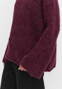 Burgundy, fluffig stickad tröja med vida ärmar och avslappnad passform, tillsammans med svarta byxor med små prickiga mönster.