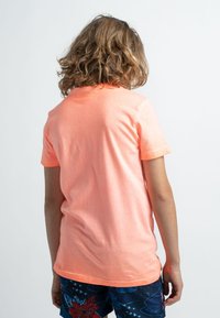 Tričko s krátkým rukávem a kulatým výstřihem v jasně korálové barvě, vyrobené z měkké bavlny, nošené s vzorovanými šortkami s modrými a květinovými motivy.