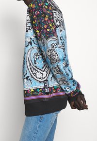Färggad sweatshirt med paisley- och blommönster, huvudsakligen blå med svarta detaljer och texturerade ärmslut; märkesdetalj i nederkant.