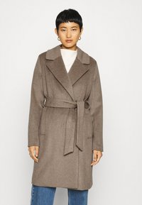 Brauner Woll-Mix-Mantel mit tailliertem Schnitt, großen Revers und langen Ärmeln. Das Model trägt einen hellen Pullover und blaue Jeans und steht vor einem schlichten Hintergrund.