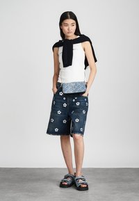 Denimshorts met witte bloemenborduring, gecombineerd met een witte tanktop en een zwarte trui die over de schouders hangt. Zwarte sandaaltjes met bandjes maken de look compleet.