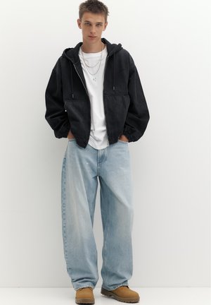 Junger Mann steht mit den Händen in den Taschen, trägt einen schwarzen Hoodie, weißes T-Shirt, hellblaue weite Jeans und braune Stiefel vor weißem Hintergrund.