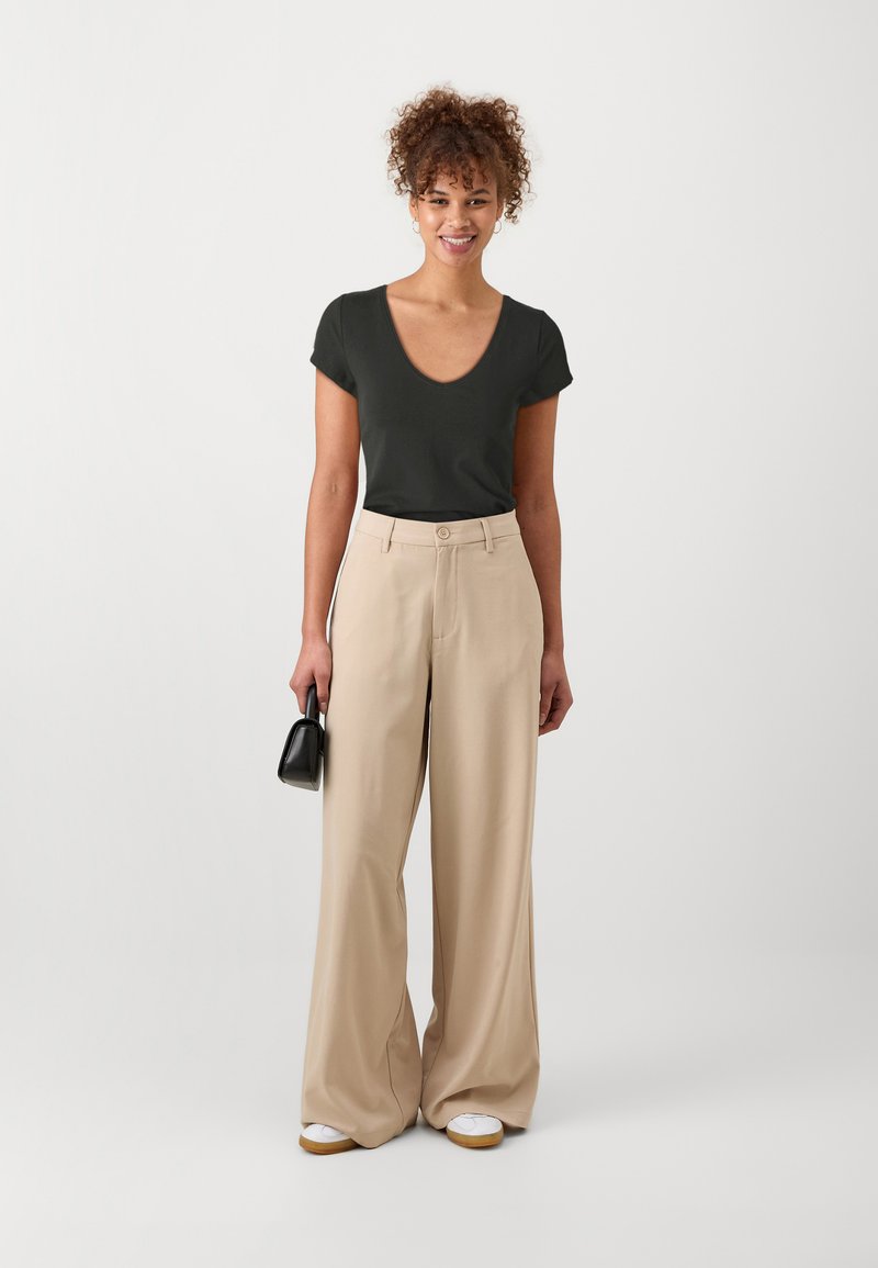 T-shirt noir à col en V, pantalon beige à jambes larges, baskets blanches, sac à main noir. Tissu lisse, coupe décontractée, et détails subtils sur le pantalon.
