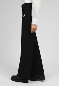 Pantalons noirs à larges jambes avec des fentes sur les côtés, ornés d'une poche décorative embellie, associés à un blouse blanche à manches longues et des bottes noires.