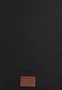 Tissu texturé noir avec un motif tissé, orné d'une étiquette en cuir marron embossée du texte "BLEND GLOBAL JEANSMAKER" en bas.