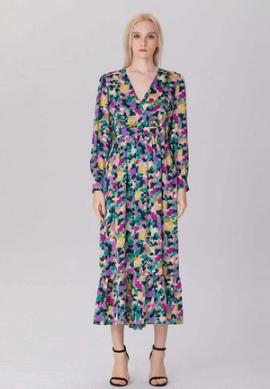 Femme debout portant une robe fleurie à manches longues et col en V avec un imprimé abstrait violet, vert, jaune et noir, associée à des talons noirs à bride cheville.