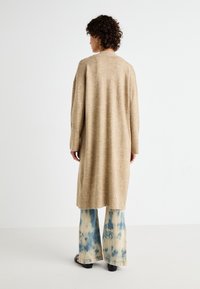 Beige lång cardigan med en texturerad stickning, med långa ärmar och en avslappnad passform, bärs över blå tie-dye vida byxor.
