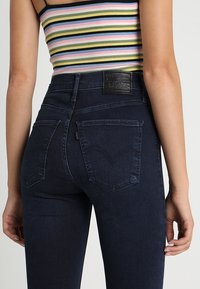 Tummansininen korkeavyötäröiset farkut, joissa on istuva malli, takataskut ja nahkainen logoplässi vyötäröllä. Sileä farkkukangas.