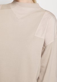 Hellbeiges Sweatshirt mit strukturiertem, geripptem Kragen, glatten Schulterpartien und minimalen Nahtdetails.