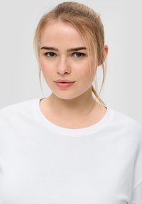 Biała bawełniana koszulka z okrągłym dekoltem, z krótkimi rękawami i gładką fakturą, noszona przez modelkę z naturalnym makijażem i prostymi włosami.