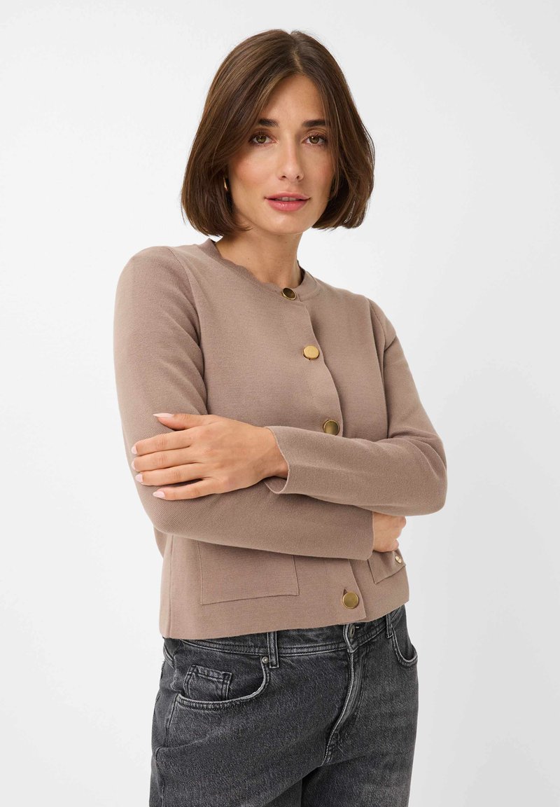 Light brown knitted cardigan with a round neck, gold-tone buttons, ribbed cuffs, and two front pockets. Paired with dark denim jeans.