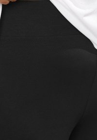 Zwarte leggings met een gladde, rekbare textuur en een hoge tailleband. De bovenrand is gedeeltelijk bedekt door een witte top.