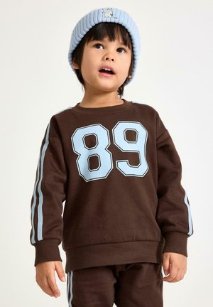 Brązowa bluza z jasnoniebieskimi akcentami i dużą liczbą „89” nadrukowaną z przodu. W zestawie pasujące brązowe spodnie i jasnoniebieska prążkowana czapka beanie.