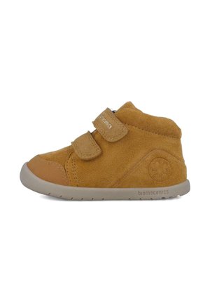 Bota de tobillo de ante marrón con dos correas de Velcro, suela de goma y logotipo grabado en el lateral. Presenta una punta redondeada y un collarín acolchado.