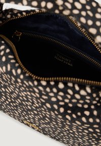 Fekete és bézs pöttyös tasak, textúrált szőrmés külső borítással, aranyszínű cipzárral és egy sötét béléssel, amelyen látható a márka címkéje.