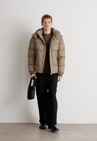 Khaki pufjakke med aftagelig hætte, lynlås, og sidelommer, parret med en brun striktrøje, sorte bukser og en sort taske.