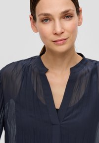 Marineblauwe blouse met een halve knoopsluiting, gemaakt van lichtgewicht doorzichtig materiaal met verticale plooien en een open decolleté. Textuurdetail op het oppervlak.