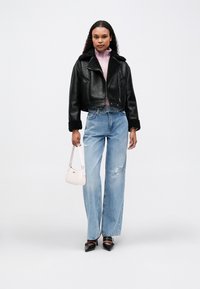 Γυναίκα με μαύρο δερμάτινο μπουφάν φλις, ροζ πλεκτό πουλόβερ, ανοιχτό μπλε σκισμένα τζιν, μαύρες μυτερές γόβες, κρατώντας μια μικρή ανοιχτό ροζ τσάντα.