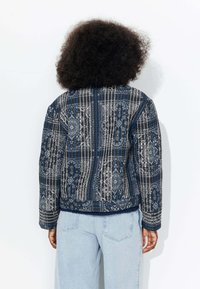 Veste matelassée avec un motif complexe bleu foncé et blanc, dotée d'un col rond et de manches longues. Tissu texturé avec des accents contrastants.