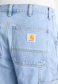 Mono de mezclilla azul claro con un gran bolsillo trasero y una etiqueta blanca que presenta un logotipo amarillo. Costuras en un amarillo contrastante.