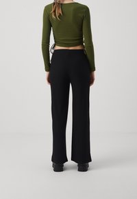 Persoon gekleed in een lange mouw olijfgroene cropped top en zwart hoge taille wijde broek, staand met de rug naar de camera tegen een effen lichte achtergrond.