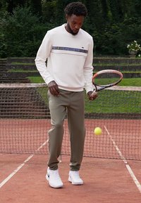 Hvit sweatshirt med en blåstripet logo, olivengrønne bukser og hvite joggesko. Tenniskølle i hånden, gul ball i luften.
