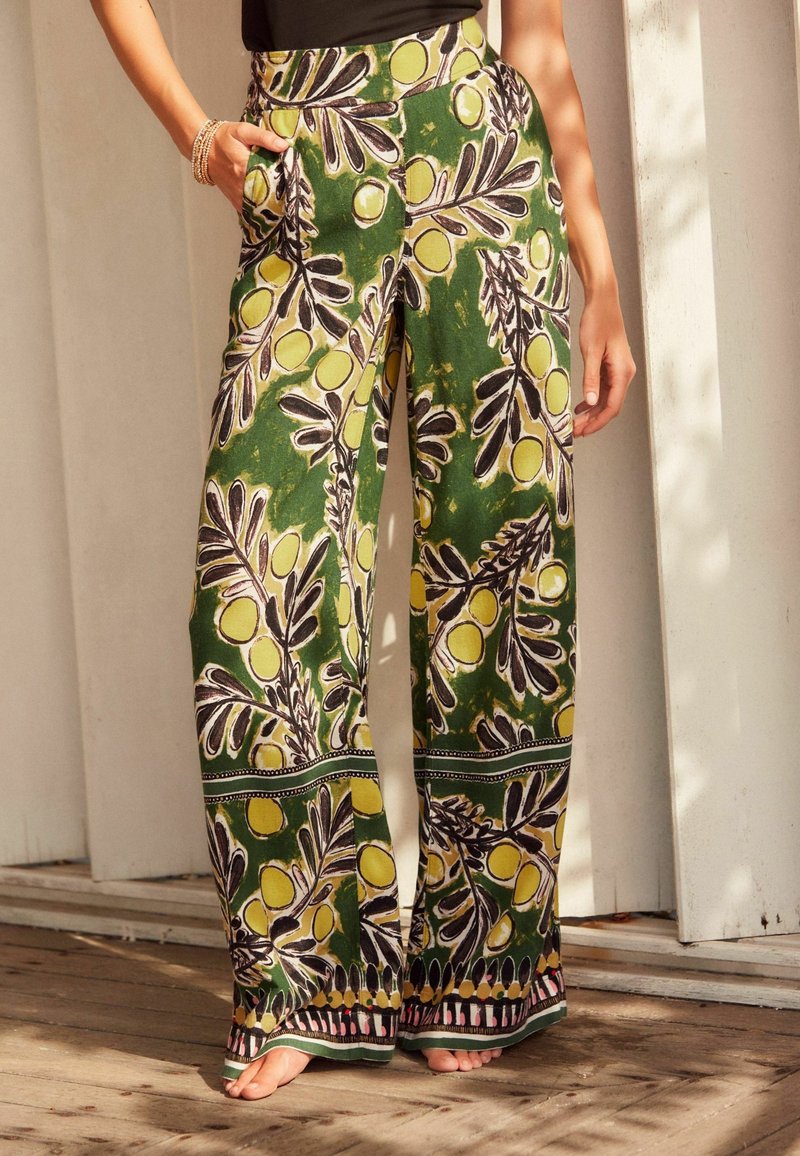 Pantalon à jambes larges avec imprimé botanique vert, jaune et noir porté par une personne pieds nus avec une main dans la poche et un haut noir.