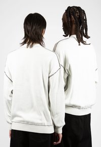 Deux personnes portent des sweat-shirts surdimensionnés gris clair avec des coutures foncées contrastantes, arborant des poignets et ourlets côtelés.