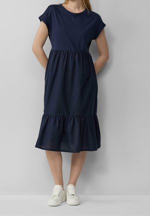 Vestido de algodón azul marino con escote redondo, mangas cortas y dobladillo con volantes en capas. Combinado con zapatillas blancas.