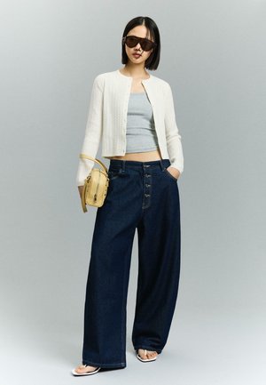 Nő fehér kardigánban, szürke crop topban, bő szárú sötét farmernadrágban, napszemüveget viselve, bézs táskát tartva, szürke háttér előtt áll.