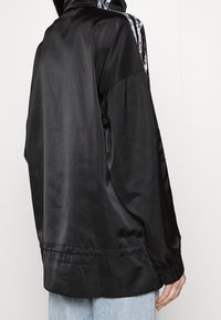 Černá saténová bunda s vysokým límcem, elastickými manžetami a květinovým zdobením na zádech. Volný střih a hladká textura.