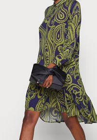 Grön och marinblå paisleymönstrad klänning med knäppkrage och långa ärmar, tillsammans med en svart satinclutch som hålls i en hand.