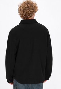 Giacca nera con colletto, realizzata in un tessuto liscio, con un fit comodo e polsini con bottoni. La parte posteriore presenta un design pulito senza motivi.