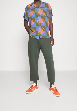 Camicia a maniche corte blu con un motivo di foglie tropicali in arancione e rosa, abbinata a pantaloni verdi e sneaker arancioni con dettagli bianchi.