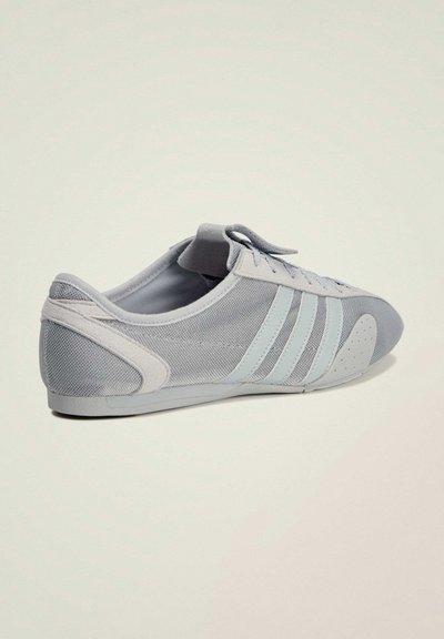 Chaussure de sport gris clair avec trois bandes pâles, détails en mesh et en daim, semelle profilée basse et lacets sur un fond uni.