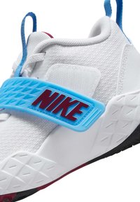 Chaussure de sport blanche avec tige en tissu texturé, sangle en caoutchouc bleue arborant le logo "NIKE" rouge, et semelle extérieure en caoutchouc noir avec un motif de crampons.