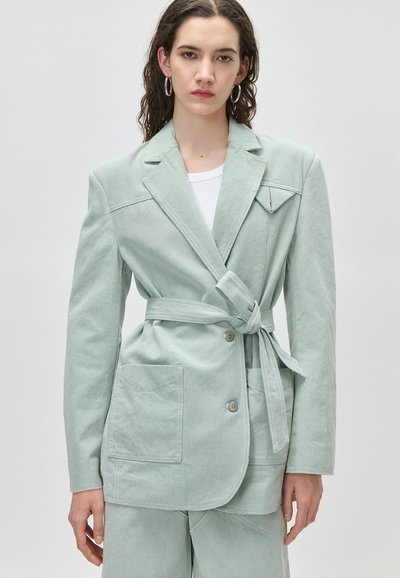 Γυναίκα που φοράει ανοιχτό πράσινο σακάκι με ζώνη, μεγάλες μπροστινές τσέπες και ταιριαστό παντελόνι, στέκεται μπροστά σε απλό φόντο.