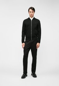 Μαύρο αθλητικό φούτερ με φερμουάρ και ραβδωτή υφή, συνδυασμένο με μαύρο παντελόνι με πλαϊνές ρίγες και μαύρα επίσημα παπούτσια.