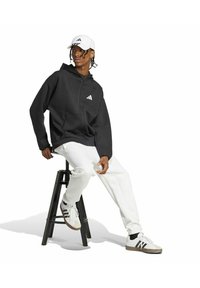 Giovane uomo in felpa nera con cappuccio, pantaloni bianchi, cappellino bianco Adidas e scarpe da ginnastica seduto su uno sgabello nero contro uno sfondo bianco.
