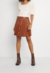 Kvinna som bär en vit stickad topp med puffärmar, en brun läderkjol med veck och framknappar, samt bruna höga ankle boots.