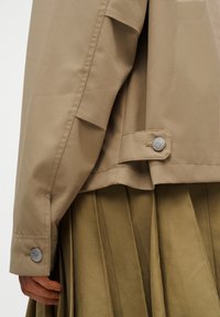 Gros plan sur une personne portant une veste beige avec un poignet boutonné et une patte réglable au-dessus d'une jupe plissée vert olive.