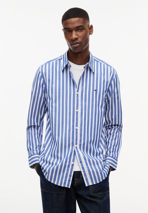 Uomo con capelli corti che indossa una camicia a righe verticali blu e bianche sopra una t-shirt bianca e jeans scuri, con le mani in tasca, che guarda davanti a sé.