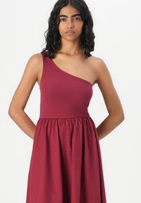 Robe bordeaux à une épaule avec un corsage ajusté en maille et une jupe évasée et froncée. Texture lisse, silhouette décontractée, sans motifs visibles.
